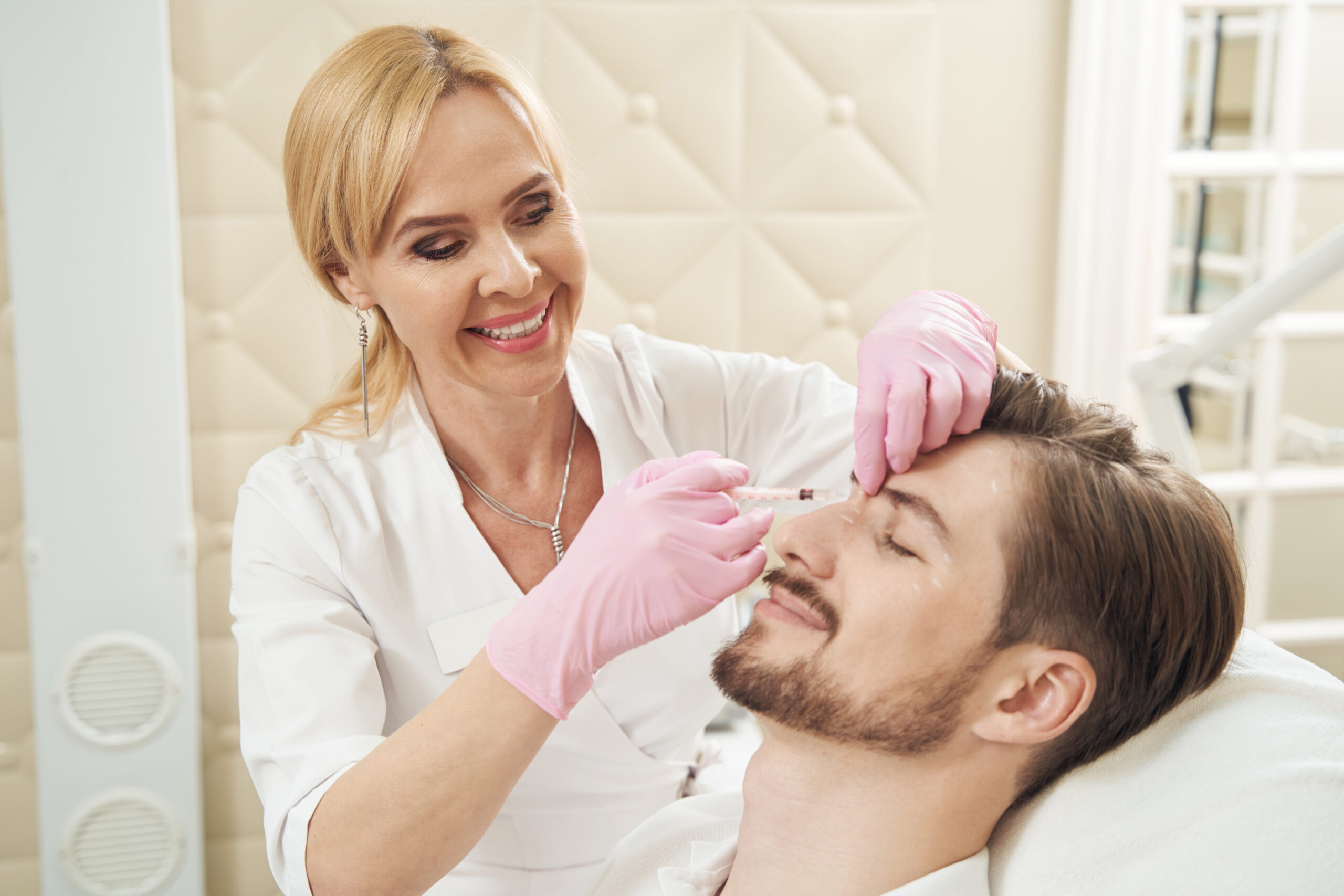 Woman smiling while injecting syringe with botulinum toxin into upper part of nose of man